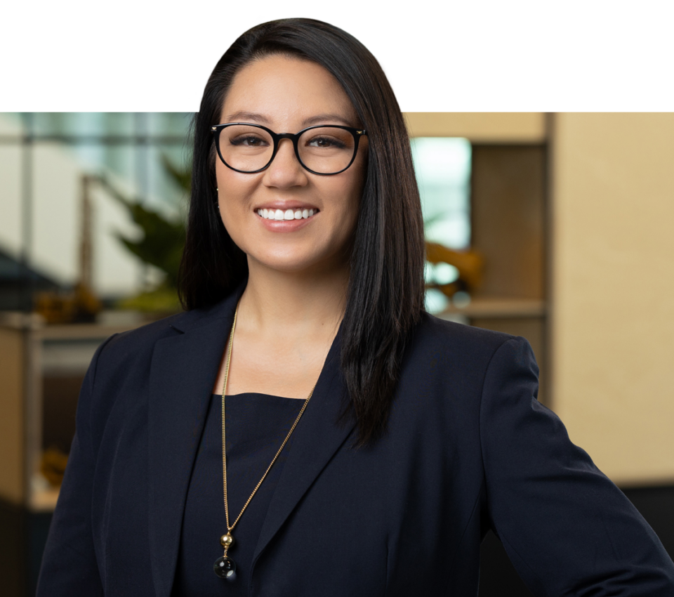 A woman with straight dark hair and glasses is smiling, wearing a navy blazer and necklace, standing in an office environment—an excellent example of modern headshots for professionals.