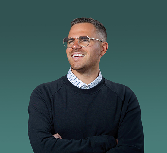 A man with short hair and glasses stands smiling with arms crossed, wearing a striped collared shirt under a dark sweater against a teal gradient background, capturing the look of modern headshots for professionals.
