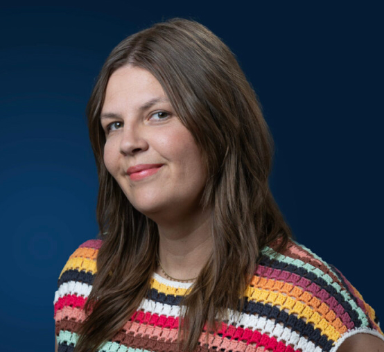 Woman with long brown hair wearing a colorful striped crocheted top, smiling slightly, posed in front of a solid dark blue background—an example of modern headshots that capture both personality and professionalism.