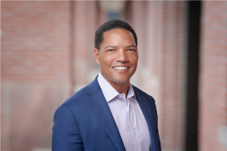 A confident man in a blue suit and light purple shirt smiles while standing outdoors in front of a blurred brick background—perfect for professional Denver headshots.