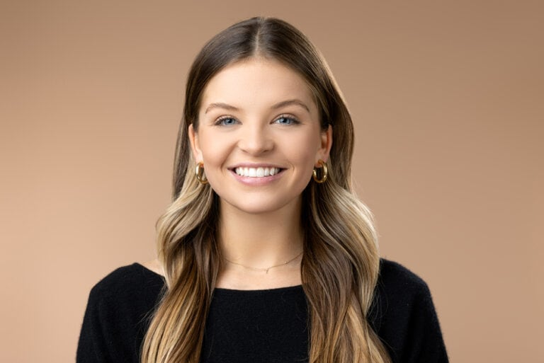 A young woman with long, light brown hair and gold hoop earrings smiles at the camera against a plain beige background. She is wearing a black top, showcasing confident photography often seen in Denver headshots.