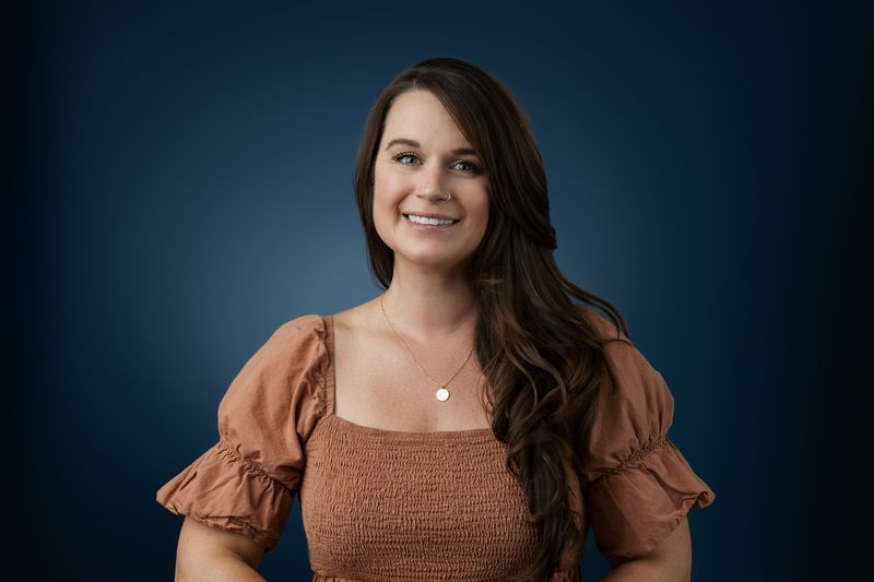 Woman with long brown hair wearing a brown puff-sleeve top and necklace, showing a natural-looking smile at the camera in this headshot against a dark blue background.