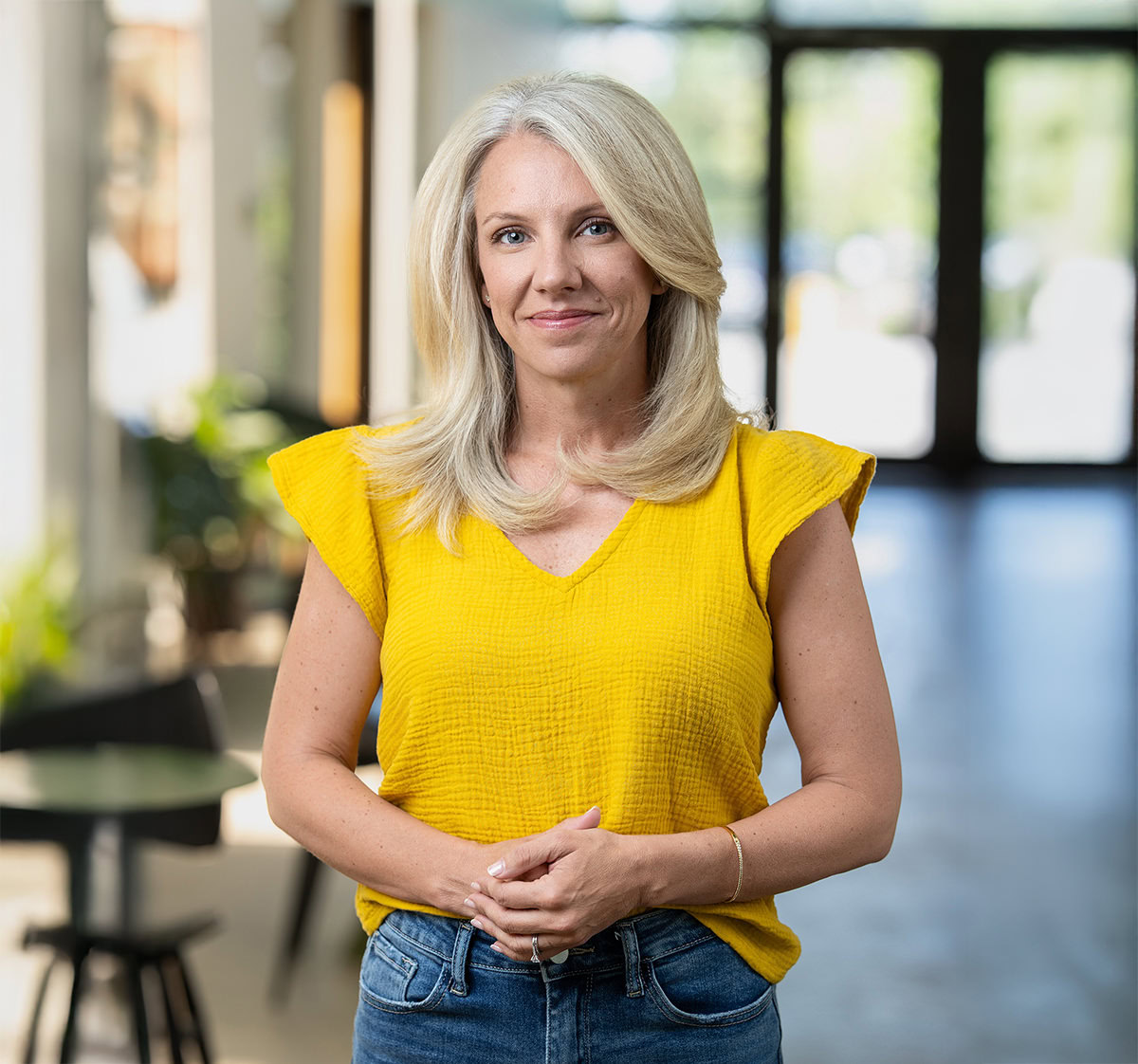 A woman with blonde hair wearing a yellow top and blue jeans stands indoors, looking at the camera with a neutral expression—perfect for individual headshots. Blurred tables, chairs, and windows fill the background.