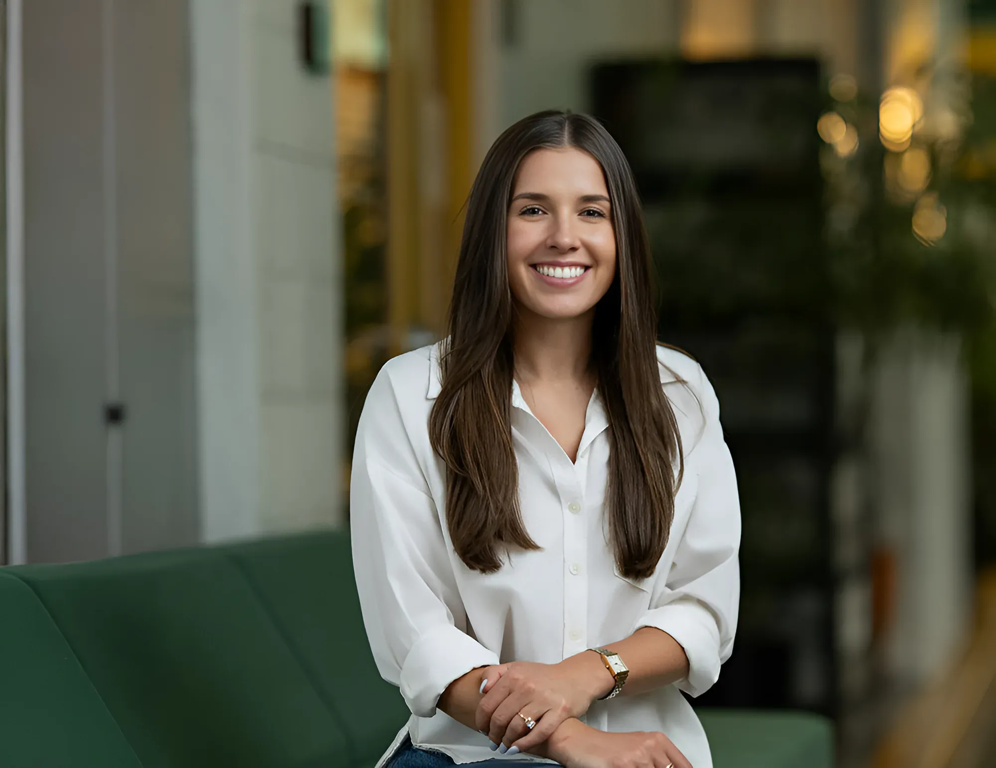 A woman with long brown hair sits on a green couch, smiling and wearing a white blouse, in a modern indoor setting—perfect for individual headshots.