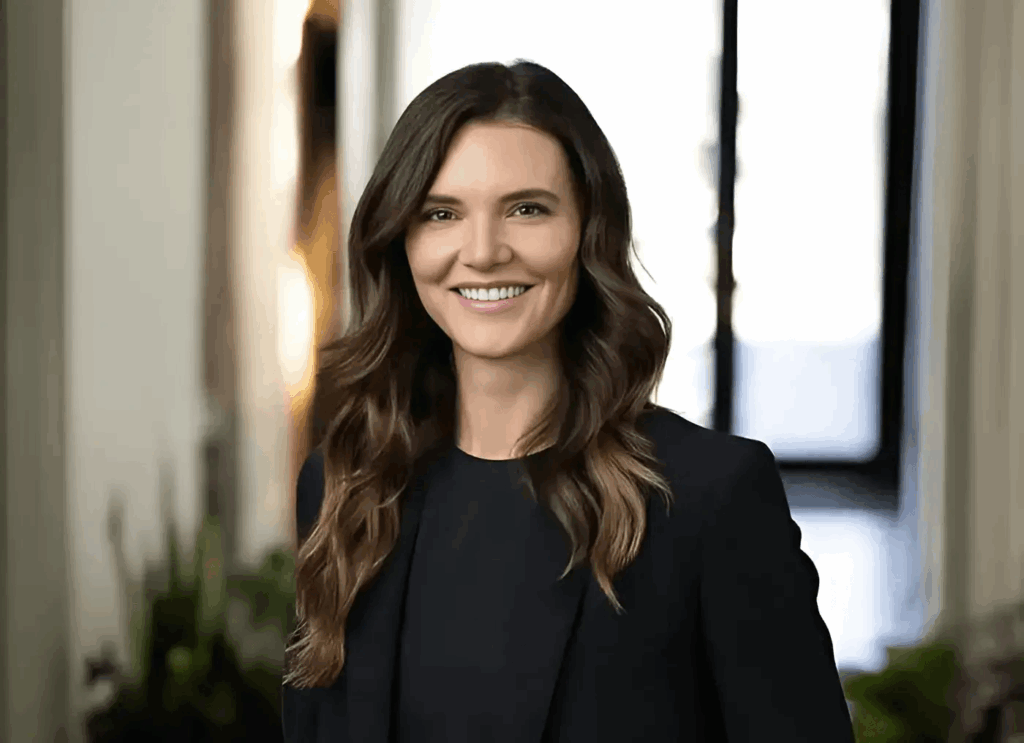 Professional woman with long wavy brown hair smiling, wearing a dark blazer.