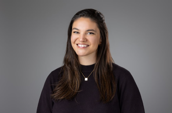 A woman with long dark hair, wearing a dark sweater and a gold necklace, smiles at the camera against a plain grey background.