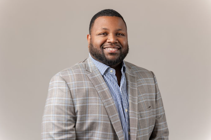 A man wearing a light plaid blazer and a striped shirt smiles at the camera against a plain light background.