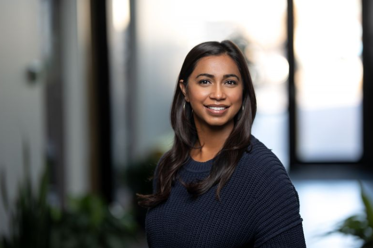 A woman with long dark hair wearing a navy sweater smiles while standing indoors in a softly lit space with blurred background.
