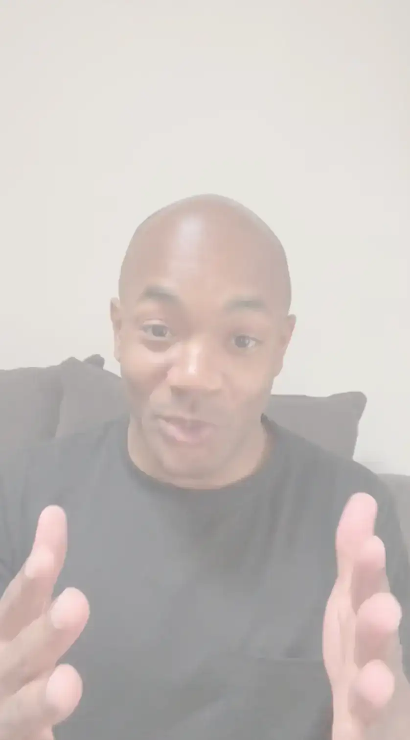 A person in a black shirt sits on a couch, facing the camera for a candid headshot with hands raised and palms open, appearing to be speaking or explaining something.