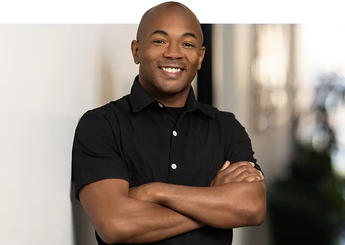 A man wearing a short-sleeve black button-up shirt stands indoors with his arms crossed, smiling at the camera in a professional headshot.
