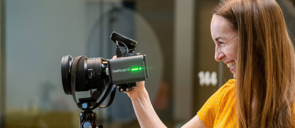 A woman in a yellow top smiles while adjusting a camera with a CamRanger device attached, mounted on a tripod.
