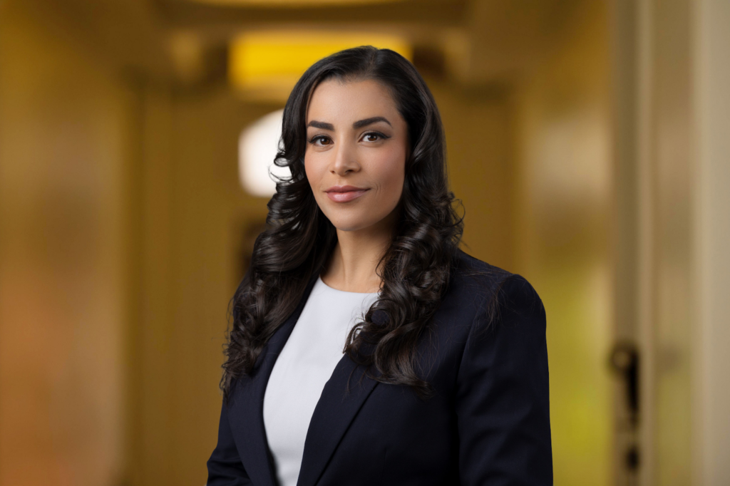 A woman with long, dark, curly hair wearing a dark blazer and white top stands in a hallway with a neutral expression.