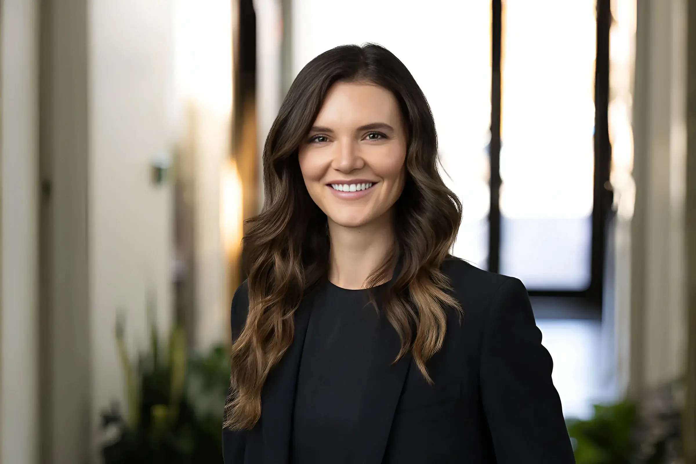 A woman with long brown hair wearing a black blazer smiles in this professional headshot, standing indoors with a blurred hallway and plants in the background.