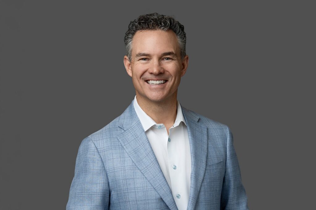 Man with curly dark hair, wearing a light blue blazer and white shirt, smiling in front of a plain gray background.