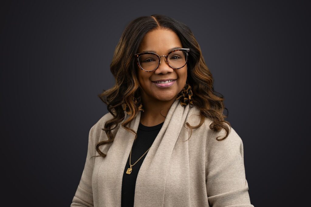 Woman with long wavy hair and glasses, wearing a beige jacket over a black top, poses against a plain dark background.