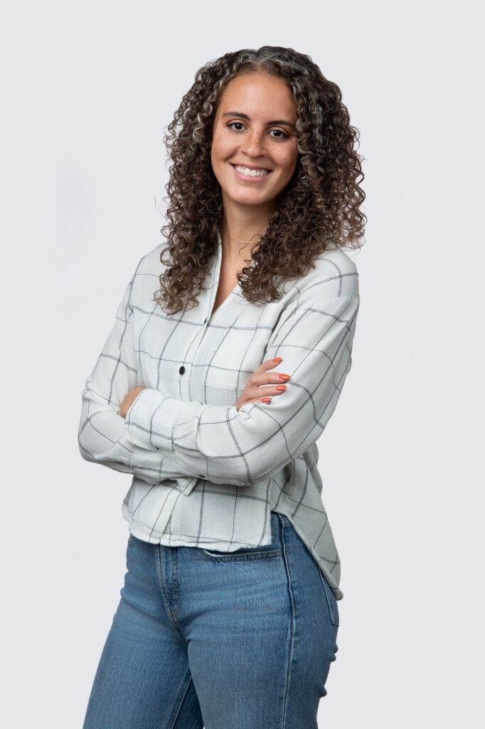 Woman with curly hair wearing a white checked shirt and blue jeans, standing with arms crossed and smiling against a plain background.