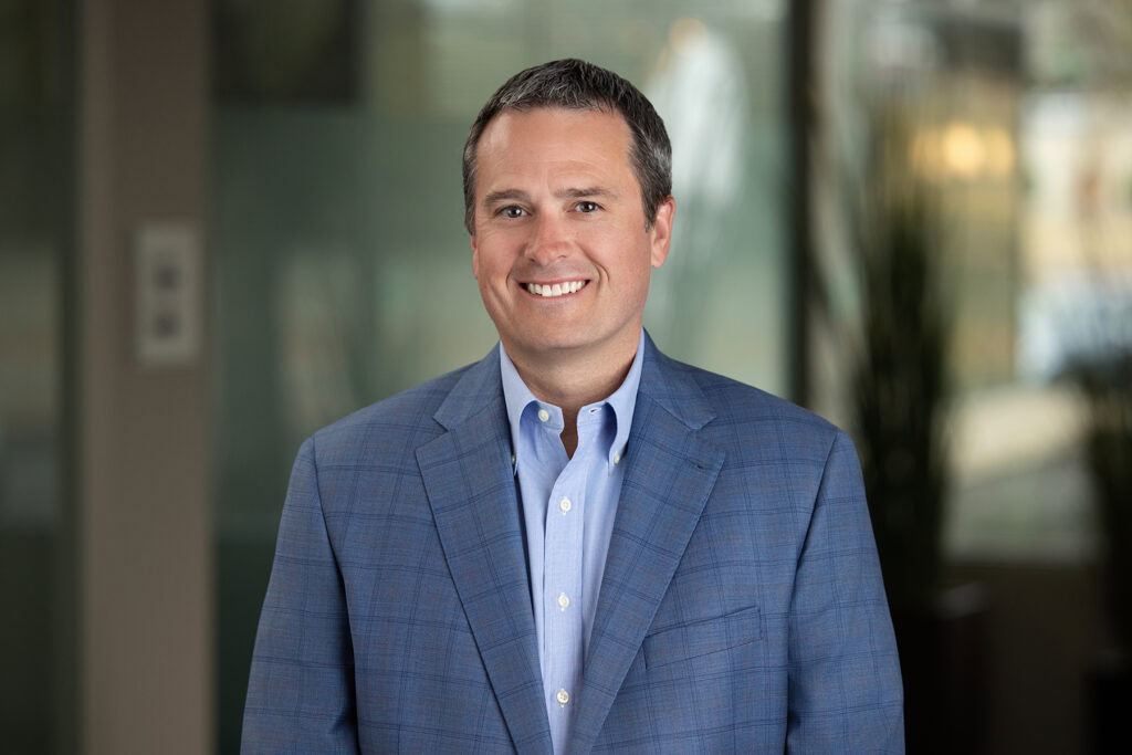 A man wearing a blue plaid suit jacket and a light blue shirt stands indoors, smiling at the camera with a blurred office background—perfect for headshots for LinkedIn or updating your CRM profiles.