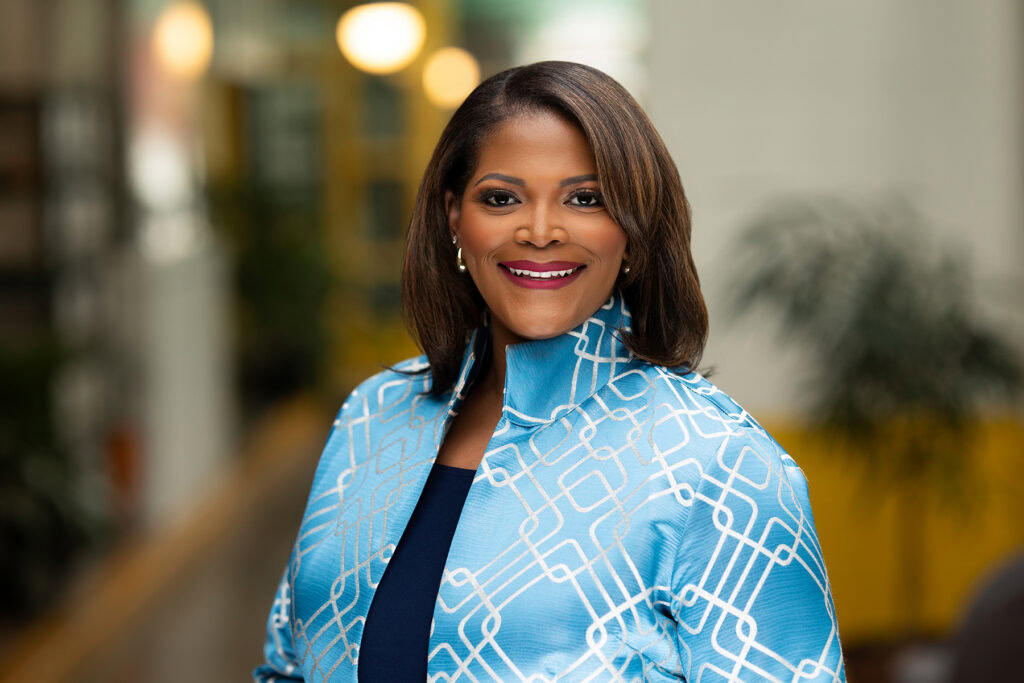 A woman with shoulder-length hair, wearing a patterned blue jacket, smiles while standing indoors with blurred plants and lights in the background—perfect for headshots for LinkedIn or updating CRM profiles.