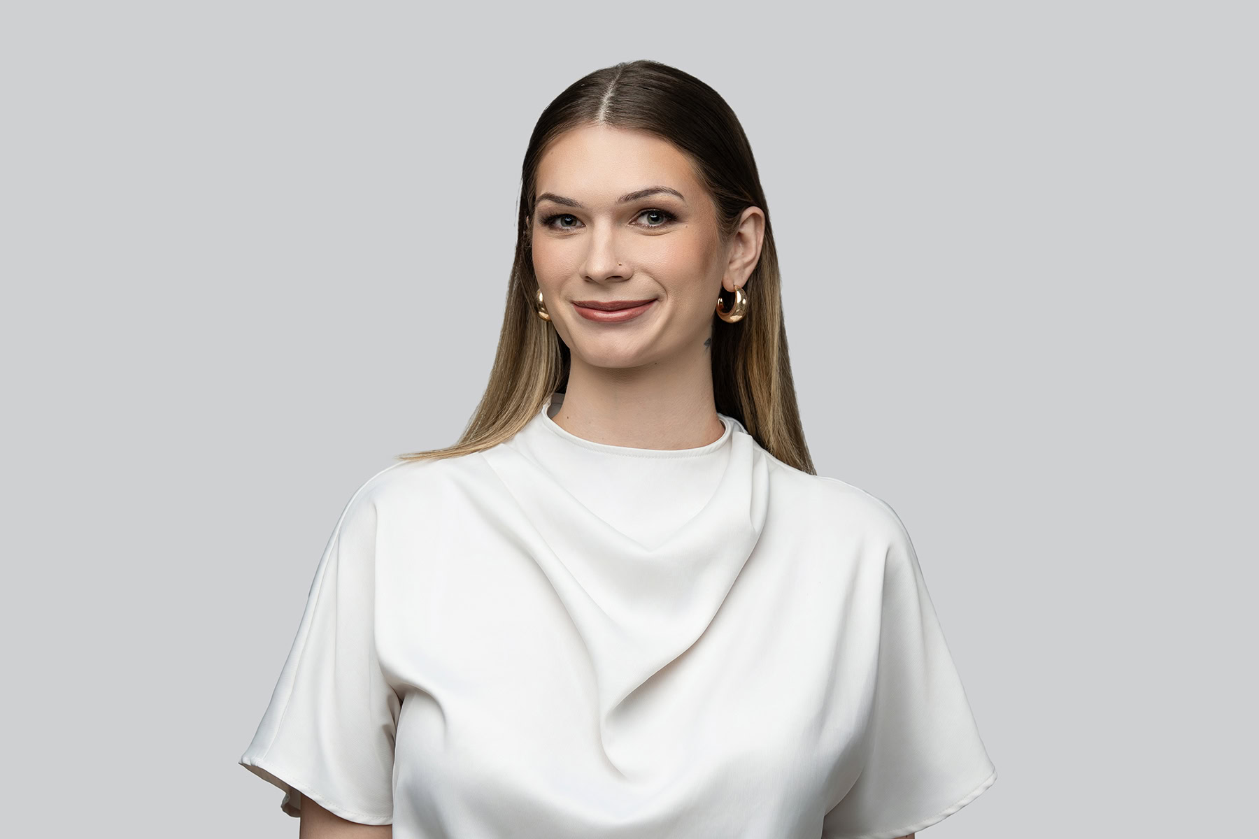 Woman with straight brown hair wearing a white blouse and hoop earrings, smiling at the camera against a plain gray background—ideal for headshots for LinkedIn or CRM profiles.